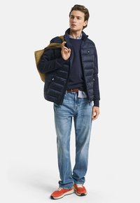 Marineblauwe puffer vest over een marineblauw trui, lichtblauw shirt, blauwe jeans en oranje sneakers. Houdt een bruine draagtas vast. Casual look.