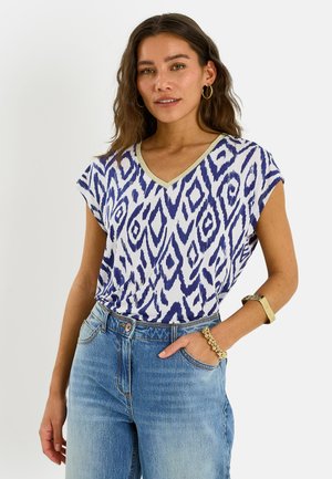 Femme aux cheveux bruns ondulés portant des boucles d'oreilles créoles, un haut bleu et blanc à motifs avec col en V, un jean bleu, un bracelet en or et une montre en or.