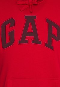 Sudadera roja con un destacado logo "GAP" en marrón oscuro en la parte frontal. El material parece suave y cuenta con un bolsillo canguro en la parte inferior.