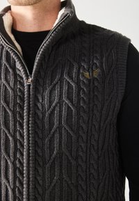 Gilet tricoté gris foncé avec un motif en relief, col haut, front zippé et doublure intérieure douce en crème. Petit logo doré sur la poitrine.