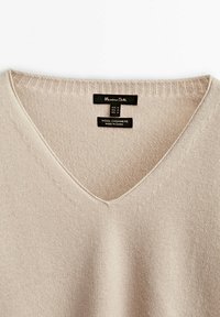 Beige wollen-kasjmier sweater met een V-hals, voorzien van een fijne brei-textuur en ribbelkraagdetails, gelabeld met verzorgingsinformatie.