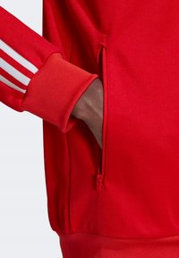 Red athletic jacket featuring a zippered side pocket, ribbed cuffs, and three white stripes along the sleeves. Smooth, textured fabric.