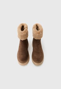 Un par de botas de invierno de ante marrón con un grueso forro de piel de tan y suelas de goma gruesa, mostradas desde arriba sobre un fondo blanco.