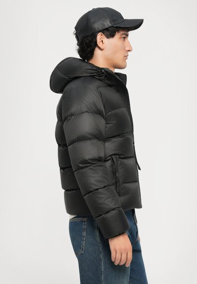 Schwarze Daunenjacke mit Kapuze, die markante gesteppte Abschnitte aufweist. Getragen über einem weißen Pullover, kombiniert mit blauen Jeans, vor einem neutralen Hintergrund.