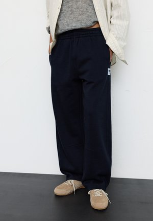 Persoon draagt losse donkerblauwe sweatpants, beige sneakers, grijze trui en een licht gestreept open overhemd, staand tegen een witte muur.