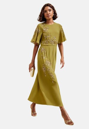 Vrouw lopend in een olijfgroene jurk met gouden bloemenborduursel, houdend een gouden clutch en draagend gouden sandalen met bandjes.