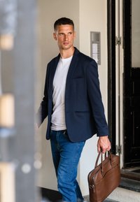 Mørkeblå blazer, hvid t-shirt, blå jeans og brun lædertaske. Slank design, glat tekstur, med detaljer af to lommer på blazeren.