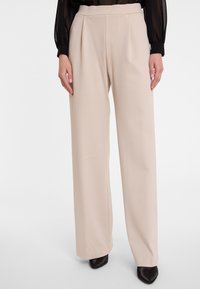 Beige wijde broek met een hoge taille en steekzakken, gemaakt van een soepele stof, gedragen met spitse zwarte schoenen.