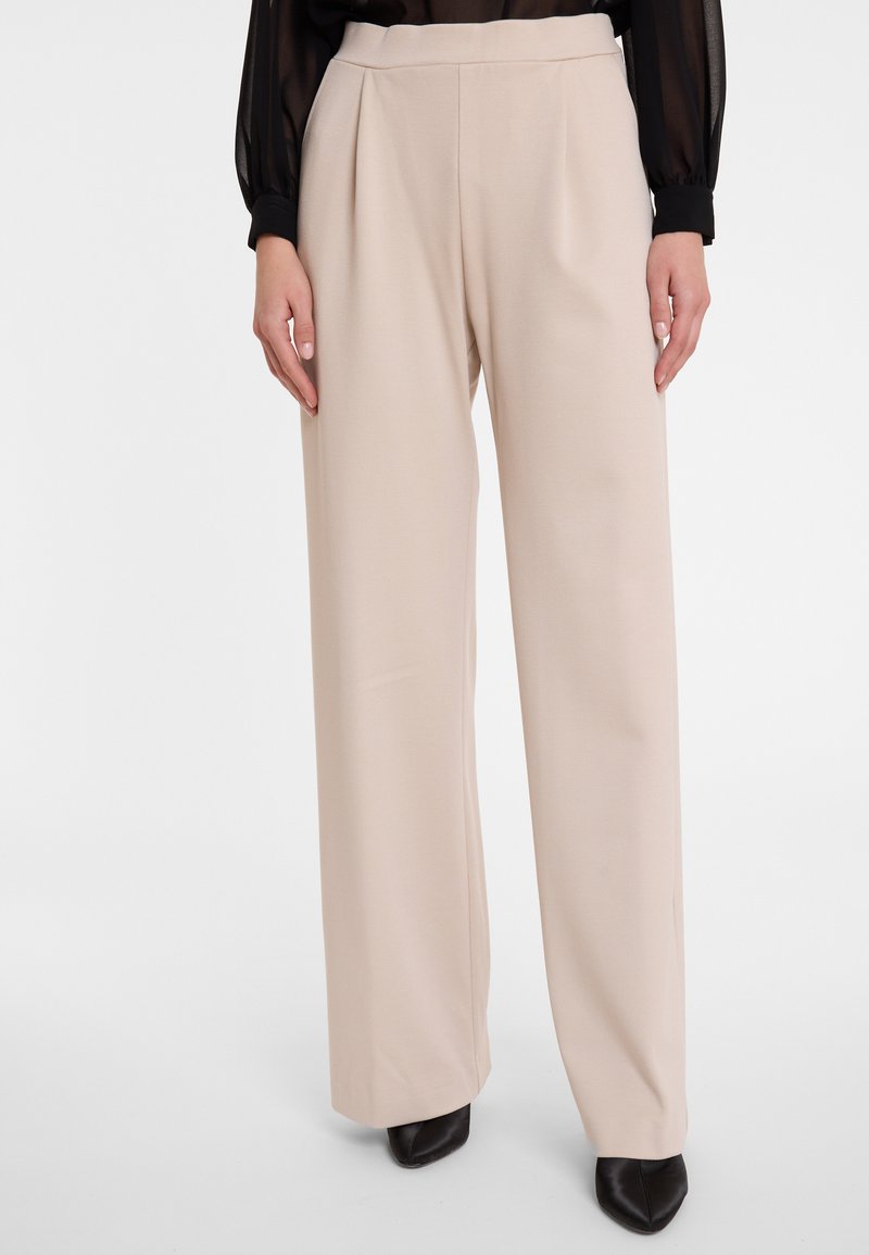 Beige wijde broek met een hoge taille en steekzakken, gemaakt van een soepele stof, gedragen met spitse zwarte schoenen.