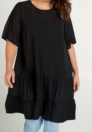 Kvinde iført en løs sort lagdelt kjole med korte ærmer og lyseblå jeans mod en ensfarvet lys baggrund.