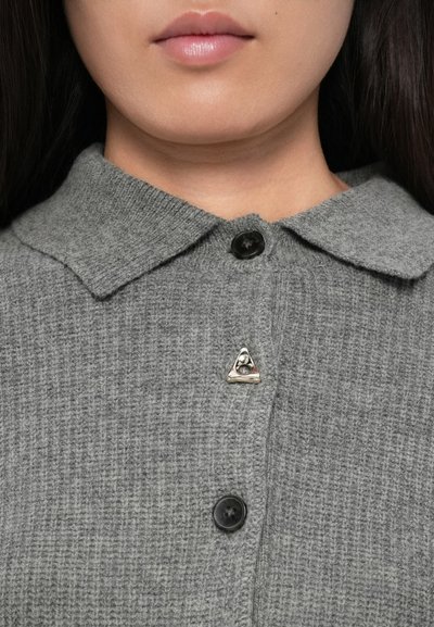 Lähedalt pildistatud inimene, kes kannab halli kudumist kašmiirkardigani mustade nööpidega, kaeluse lähedal on kolmnurkne hõbedane rinnamärk.