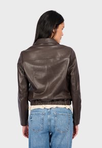 Veste en cuir marron au design ajusté, avec un col et un ourlet élastiqué. Portée par-dessus un jean bleu. Texture subtile visible.