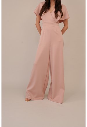 Kvinde iført en rosa buksedragt med vidde ben, korte flagermusærmer og en V-hals med slå-om front, stående med hænderne bag ryggen.