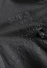 Des gouttes d'eau couvrent un tissu noir Superdry avec une fermeture éclair visible sur le côté droit.