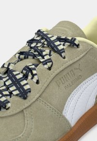 Gros plan d'une basket beige Puma Palermo avec des lacets noirs et blancs à motifs et une bande en cuir blanc sur le côté.