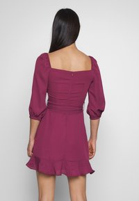 Kort lila klänning med puffärmar, fyrkantig halsringning och volangkant. Tillverkad av lätt tyg, den har en figursydd byst och dragkedjestängning.