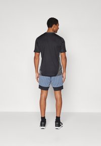 Schwarzes Sportshirt mit kurzen Ärmeln und rundem Halsausschnitt. Kombiniert mit hellblauen Shorts mit einem dunkelblauen Saum. Schwarze Turnschuhe.
