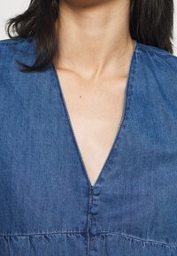 Denimová šaty s výstřihem do V, předními knoflíky a nařaseným pasem. Materiál je strukturovaný a barva je středně modrý ombré.