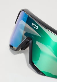 Black sports sunglasses with a large, wraparound design. They feature gradient green-blue lenses and textured side accents. Logo visible.