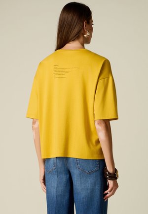 Vrouw met lang haar die een geel T-shirt met korte mouwen en kleine tekst op de rug draagt, blauwe spijkerbroek, creolen, en armbanden, van achteren gezien.
