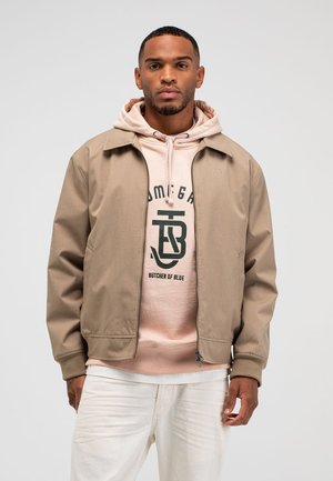 Man draagt een beige jack met ritssluiting over een lichtroze hoodie met grafische tekst, witte broek, staand tegen een effen lichte achtergrond.