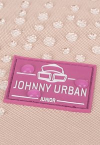 Rózsaszín textúrált háttér kiemelkedő átlátszó pontokkal. Egy lila gumicímke fehér szöveget tartalmaz: "JOHNNY URBAN JUNIOR" és szemüvegeket ábrázoló ikonokat.
