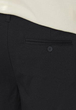 Pantalons noirs en tissu tissé, dotés d'une poche arrière avec une ouverture horizontale et une fermeture à bouton, associés à un haut de couleur claire.