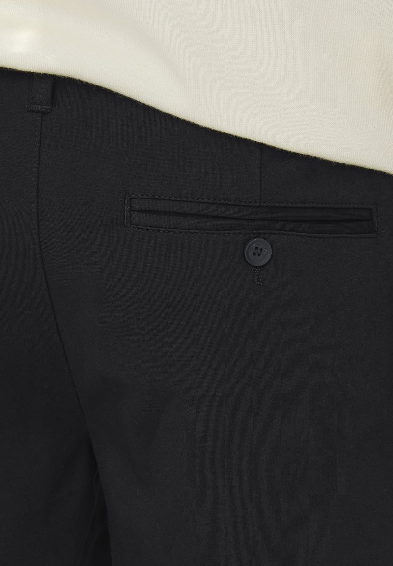 Pantalons noirs en tissu tissé, dotés d'une poche arrière avec une ouverture horizontale et une fermeture à bouton, associés à un haut de couleur claire.