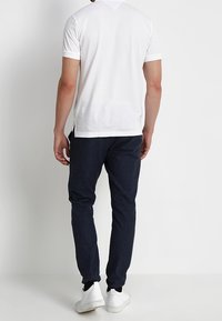 T-shirt blanc à manches courtes avec un col en V, associé à un pantalon bleu foncé et des baskets blanches, vu de dos sur un fond uni.