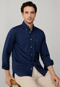 Chemise bleue marine à boutons avec un col pointu, manches longues retroussées aux poignets, poche poitrine simple, et un tissu lisse et texturé.