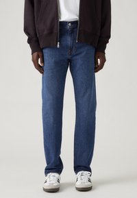 Homme portant un jean droit bleu, des baskets blanches avec des rayures noires, une chemise blanche et une veste noire zippée, debout devant un fond uni.