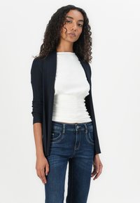 Navy ribbed open-front cardigan with long sleeves paired with a white ruched top and dark denim jeans. Smooth texture and fitted design.