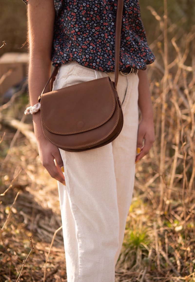 Borsa a tracolla in pelle marrone con patta arrotondata, dettagli di cuciture visibili, indossata con pantaloni di velluto a coste color crema in un ambiente naturale.