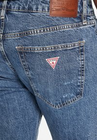 Jeans in denim di colore azzurro chiaro, con una finitura testurizzata, design classico a cinque tasche e una toppa triangolare rossa e bianca sulla tasca posteriore.