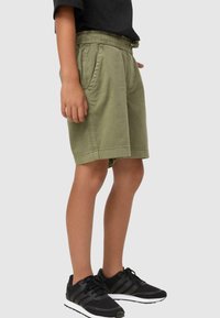 Shorts vert olive avec une taille élastique, poches latérales et coupe décontractée. Portés avec des baskets de sport noires. Tissu en coton lisse.