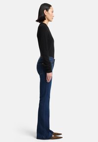 Schwarzer Pullover, dunkelblaue ausgestellte Jeans, spitze Schuhe mit Leopardenmuster. Seitenansicht, die den Sitz und die Form des Outfits zeigt.