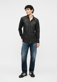 Mladi moški stoji, oblečen v črno jeans srajco, izprane modre kavbojke in črne superge z belimi črtami, gleda v stran.