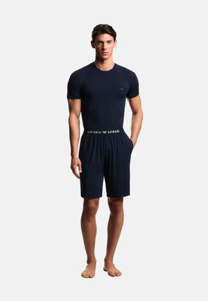 Giovane uomo in piedi a piedi nudi che indossa una maglietta nera aderente e pantaloncini neri con cinturino con logo Emporio Armani.