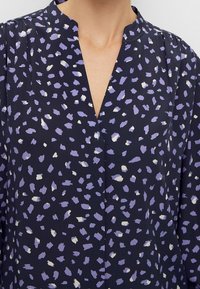 Marinebluse mit V-Ausschnitt, die ein Muster aus lavendelfarbenen und weißen Pinselstrichformen präsentiert. Leichter, glatter Stoff mit einem entspannten Schnitt.