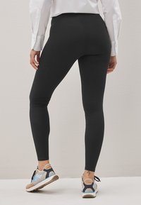 Personne portant des leggings noirs et des baskets blanches avec des accents bleus et beiges, debout devant un fond clair uni, vue de dos.