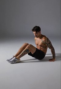 Szürke sportcipők narancssárga részletekkel, fekete rövidnadrág és karokon látható tetoválások. A téma egy világosszürke felületen ül.