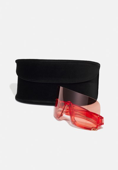 Gafas de sol rojas transparentes con un gran diseño envolvente, que presentan acentos dorados en las patillas. Acompañadas de una funda blanda negra.