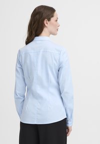 Svijetloplava košulja dugih rukava s krojenim fitom i glatkom teksturom. Ima klasični ovratnik i detalj na leđima.