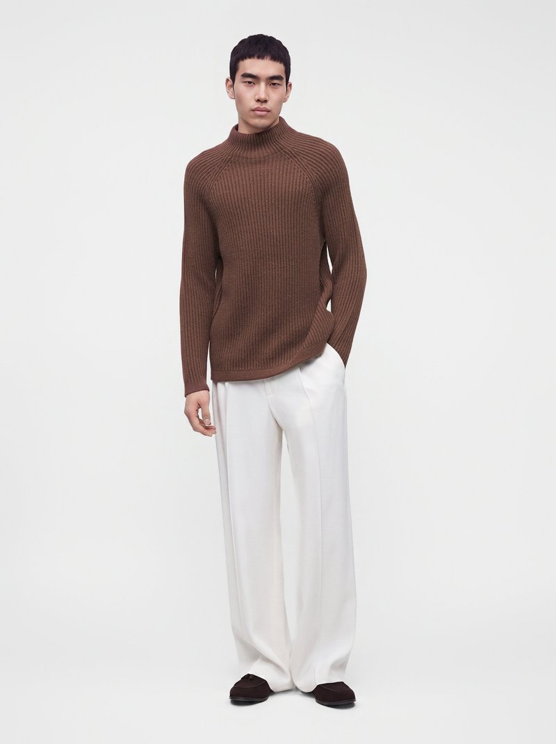 Jeune homme portant un pull roulé côtelé marron, un pantalon blanc à jambes larges et des chaussures marron foncé, debout devant un fond blanc uni.