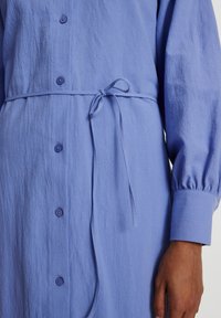 Hellblaues Hemdkleid aus strukturiertem Stoff, mit durchgehender Knopfleiste, langen Puffärmeln und einer gebundenen Taillenpartie zur Betonung der Silhouette.