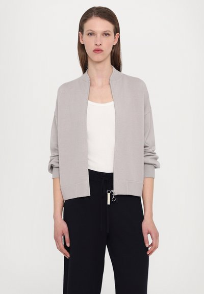 Svetlo siva skrajšana jakna z zadrgo in stoječim ovratnikom, rebrastimi manšeti in robom, v kombinaciji z belo majico brez rokavov in temnimi hlačami.