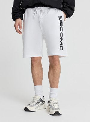 Mężczyzna noszący białe szorty z troczkiem z napisem "BECOME" na lewej nodze, białe skarpety oraz białe i czarne buty do biegania.