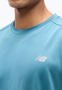 Κοντινή λήψη ενός ατόμου που φοράει φωτεινό γαλάζιο ριμπαριστό αθλητικό μπλουζάκι με λευκό λογότυπο New Balance στο στήθος.