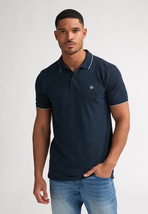 Mann mit kurzen Haaren, der ein marineblau gestreiftes Polohemd und hellblaue Jeans trägt, steht mit einer Hand in der Tasche.