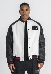 Læder- og tekstil-varsityjakke, hvid krop, sorte ærmer, ribbede røde og sorte detaljer, med et logo på brystet og trykknapper.